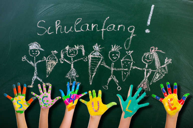Tafel mit Schriftzug Schulanfang und Zeichnung Kinder mit Schultueten davor bunte Kinderhaende